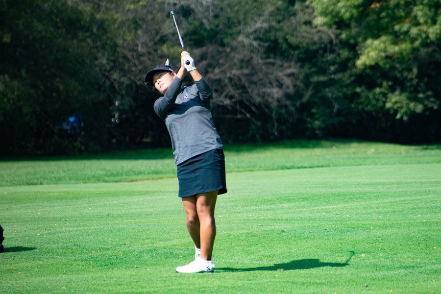 A girl with black hair in a gray shirt and black skirt swings a silver golf club on a green golf course.