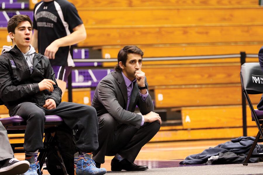 Wrestling coach in suit squats on sideline.