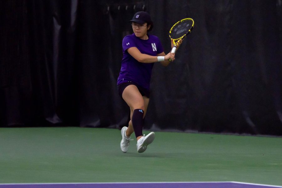 A tennis player in purple holds a racket with two hands, about to hit a ball.