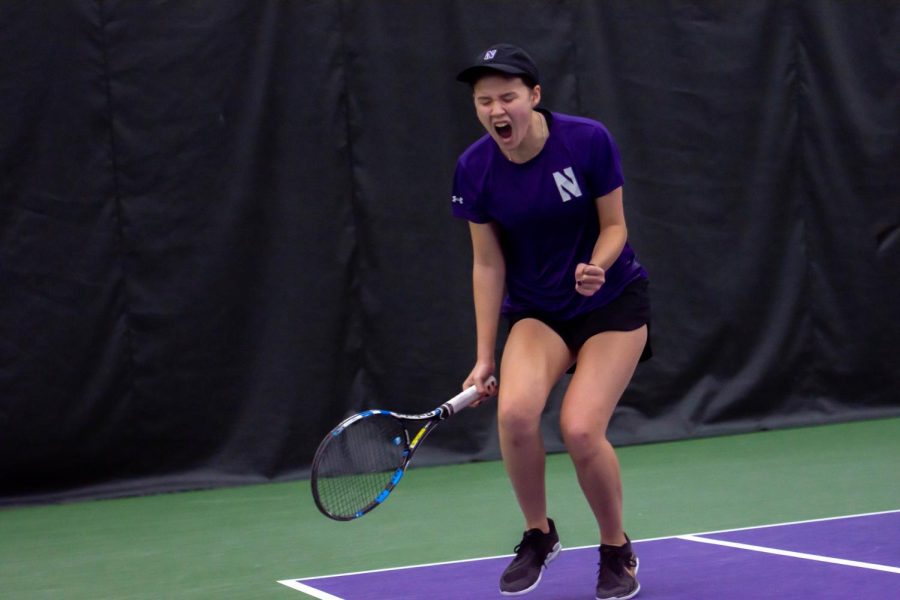 Tennis player in a purple shirt holds a racket with fist pumped and excited look on her face.