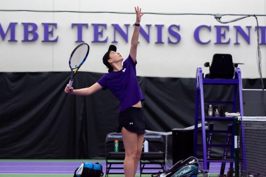A tennis player in a purple shirt has her hand extended in the air as she is about to hit a serve.