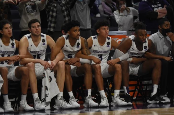 Five basketball players in white jerseys and white shorts lock arms on the bench.