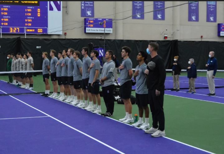 Tennis players in gray shirts and black shorts line up on the side of a tennis court with hands over their hearts.