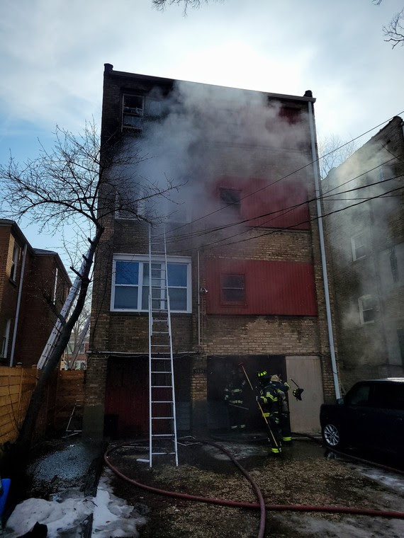 Smoke+billows+out+of+a+tall+building.+A+ladder+reaches+up+to+one+of+the+windows%2C+and+firefighters+stand+near+the+entrance.