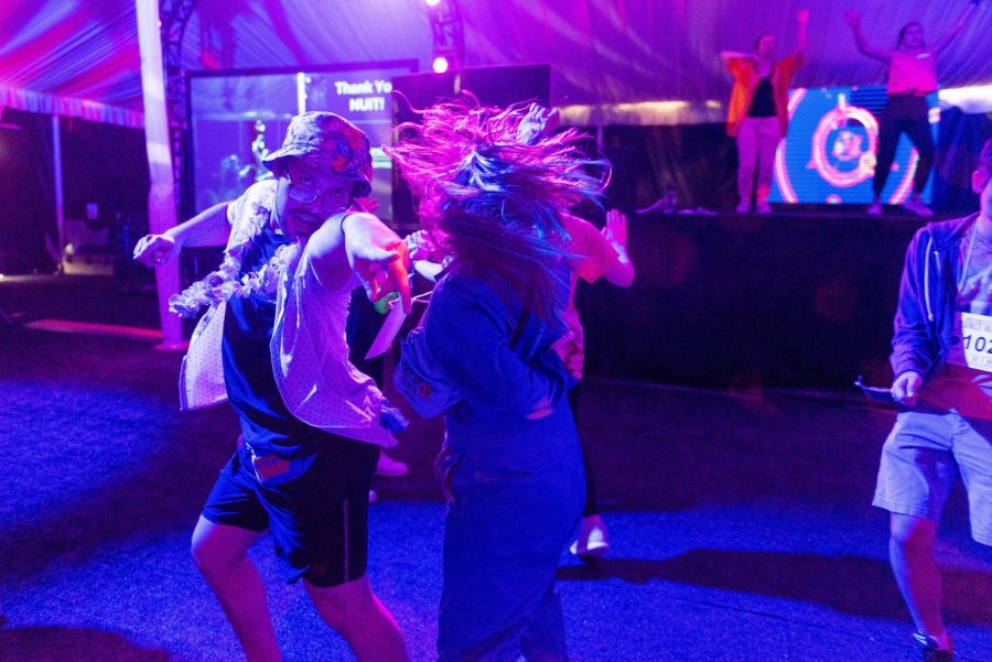 A man in a bucket hat points at the camera and a girl whips her hair. They are under blue and pink lights.