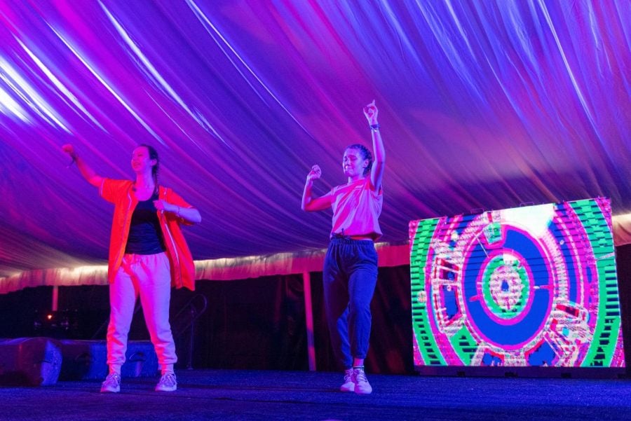 Two women illuminated with pink lights dance on a stage. There is a screen behind them with a colorful graphic.