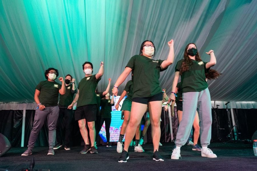 A group of people wearing green shirts dance under green lights on a stage. 