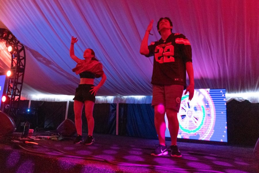Two people dance on a stage with pink and blue lights. They have one one arm up and one arm down.