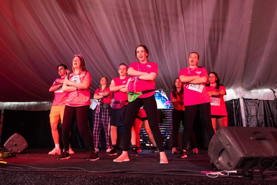 A group of about nine people wearing pink shirts dance on stage under pink lights.Their hands are all crossed. 