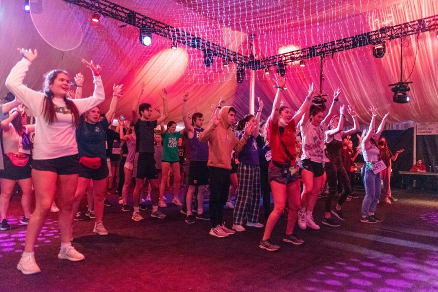 A large group of people dance in lines under pink lights. All of their hands are stretched above their heads. 