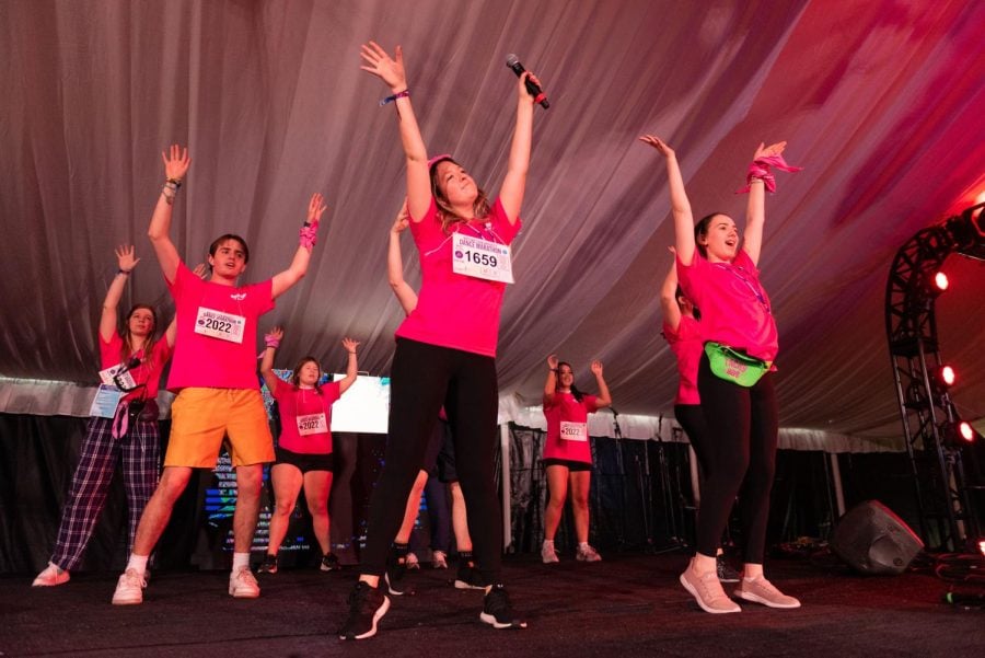 A group of dancers in pink shirts dance on a stage. Both of their arms are stretched toward the ceiling. 