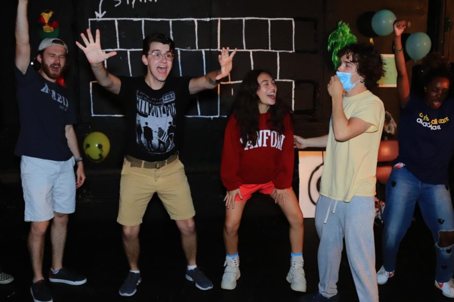 Five actors pose animatedly in front of black backdrop with white boxes drawn on. Balloons and other props are visible in the background.