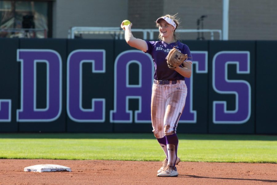 Softball player in purple-and-white uniform holding a ball in right arm stands on dirt infield in front of dark-colored wall.