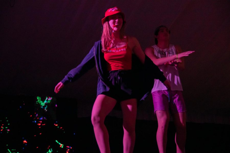 A student in a red shirt and red hat dances on stage. Another student dances behind them.