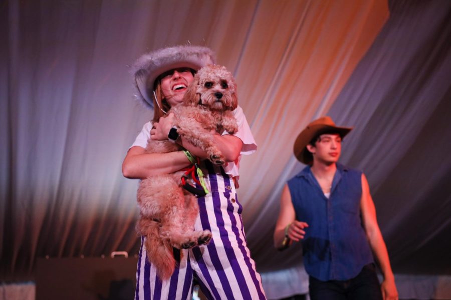 A girl in striped overalls and white cowboy hat holds a dog.
