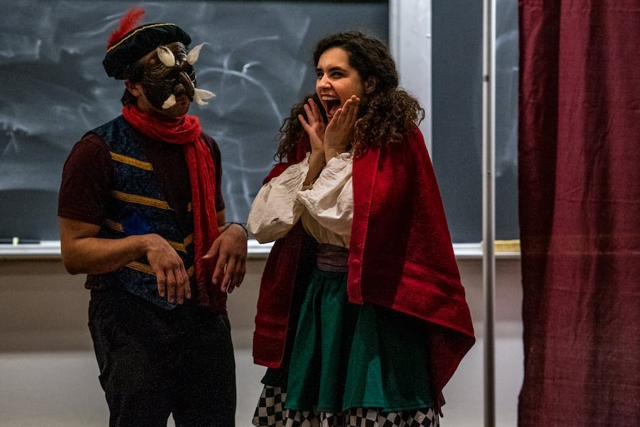 One actor in a skirt, blouse and red cape holds their hands to their face in delight. Another in a vest, scarf and plague-era mask watches them with their hands limp in front of them.