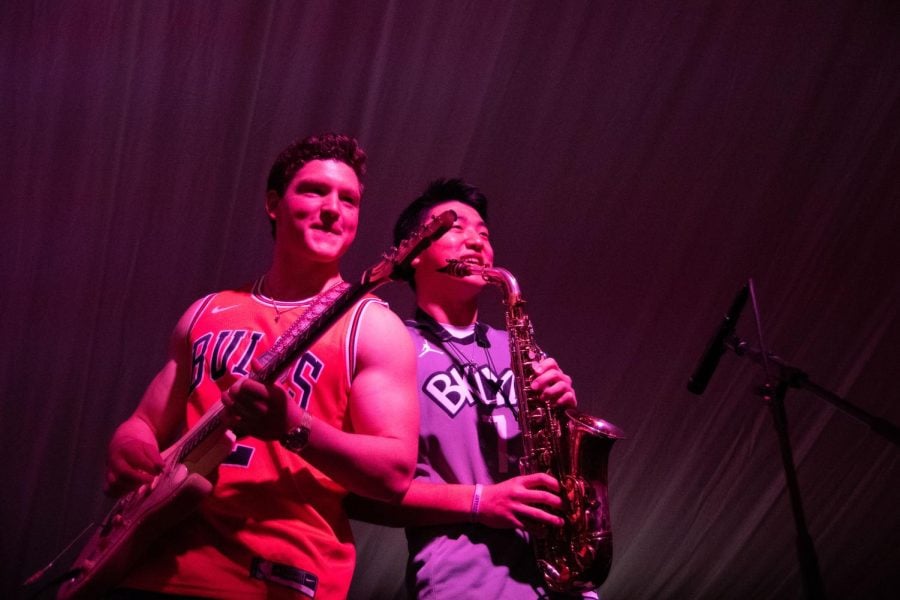 Two morning Dew Band members wearing basketball jerseys lean against each other when performing. The man on the right is playing the bass while the man on the right is playing the saxophone.