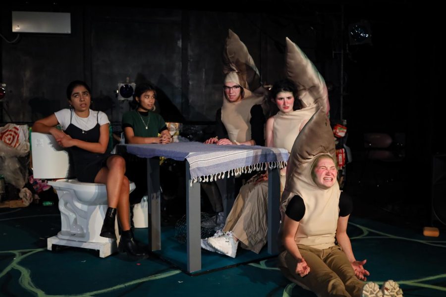 Five Vertigo Productions cast members onstage. Two girls on the left wear casual clothing while sitting at a table. The three on the right wear trout costumes.