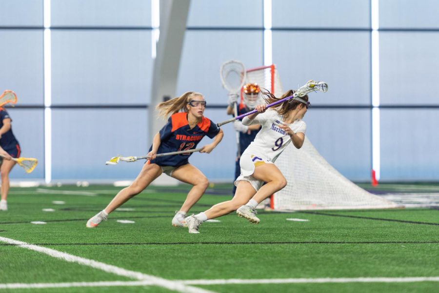 A woman in a white jersey bolts away from someone in a blue jersey. There is a ball in her lacrosse stick.