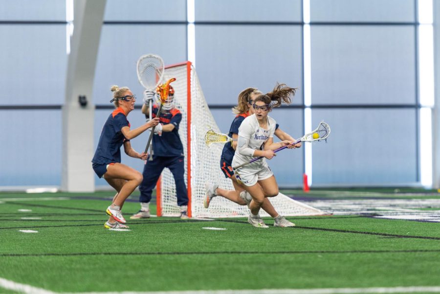 A woman in a white jersey plays lacrosse. She is surrounded by three players in blue jerseys.