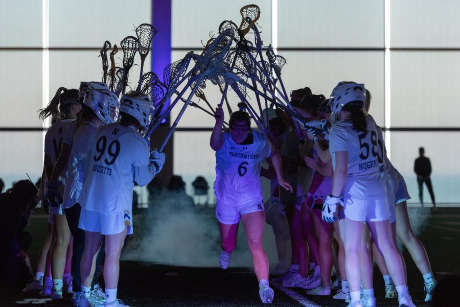 A woman in a white jersey runs under a tunnel of lacrosse sticks while fog spreads out behind her.