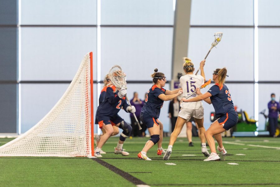 A woman in a #15 white jersey shoots a goal toward a lacrosse net.