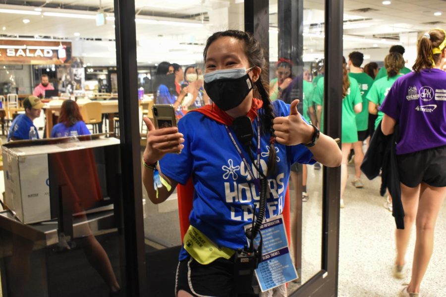 A person with a blue shirt, red cape and walkie talkie giving the thumbs up, standing in the doorway with people exiting in the background. 