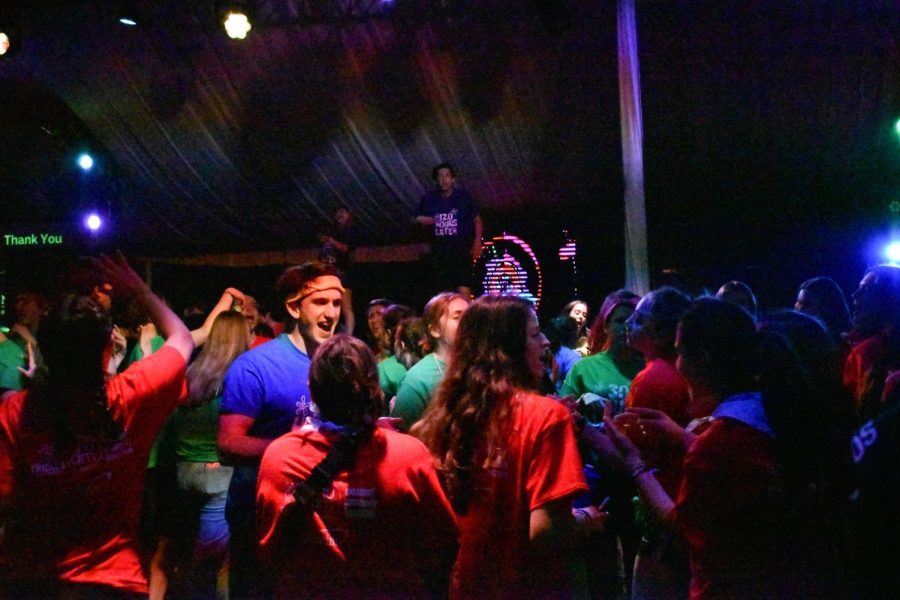 A crowd of people in red, green and blue shirts in front of a stage. On the stage is a person leaning forward and a screen with colorful lights on it.