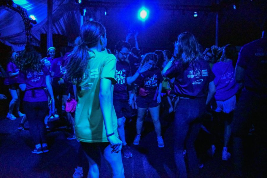 A crowd of people dancing in blue lighting. The dancer in the front wears a green shirt and the rest are wearing blue shirts. 
