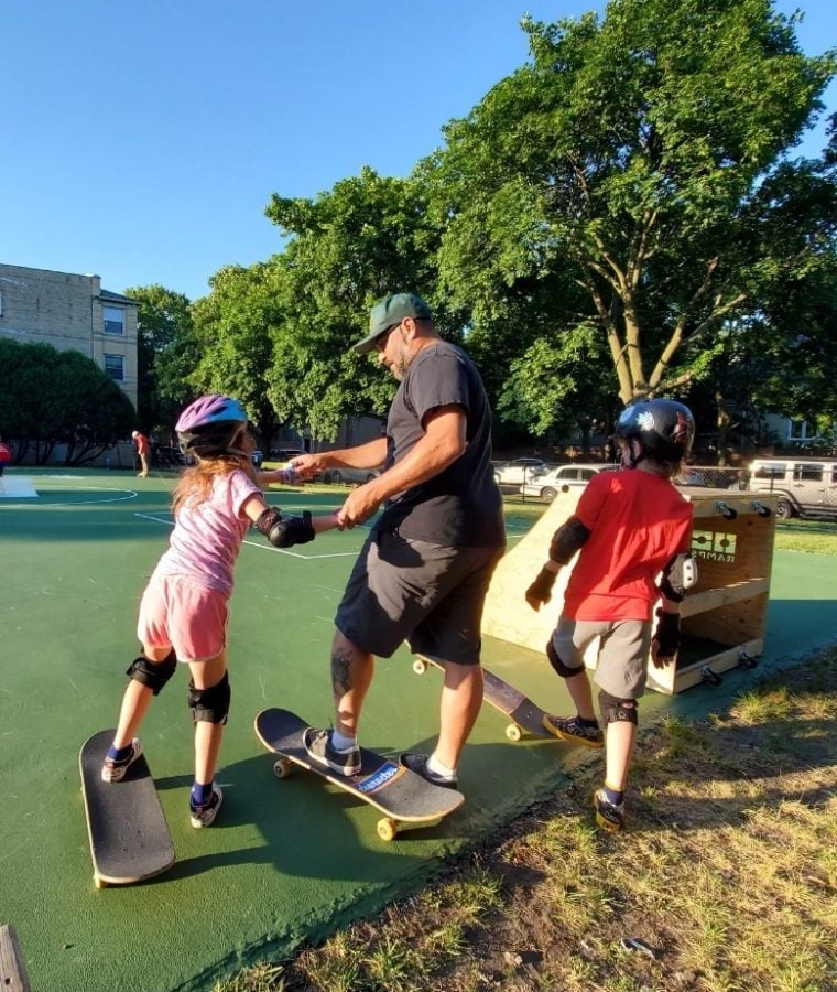 Ald. Juan Geracaris (9th) and his two children practice skateboarding. There are green trees in the background.