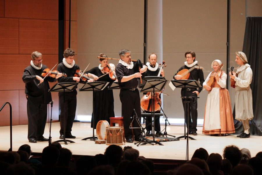 Seven musicians in historical dress stand on stage with various instruments.
