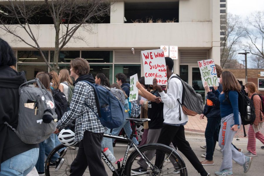 A group of students walk on the street. Some hold signs reading phrases such as “We Want a Sustainability Coordinator. One walks with a bicycle.