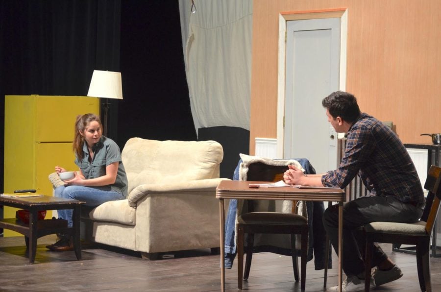 Two people sit on opposite sides of a set. In the background are a yellow fridge, white lamp, black curtain and a gray door.
