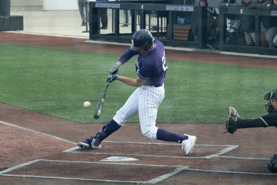 Baseball player in purple shirt and white pants swings their bat at the baseball.