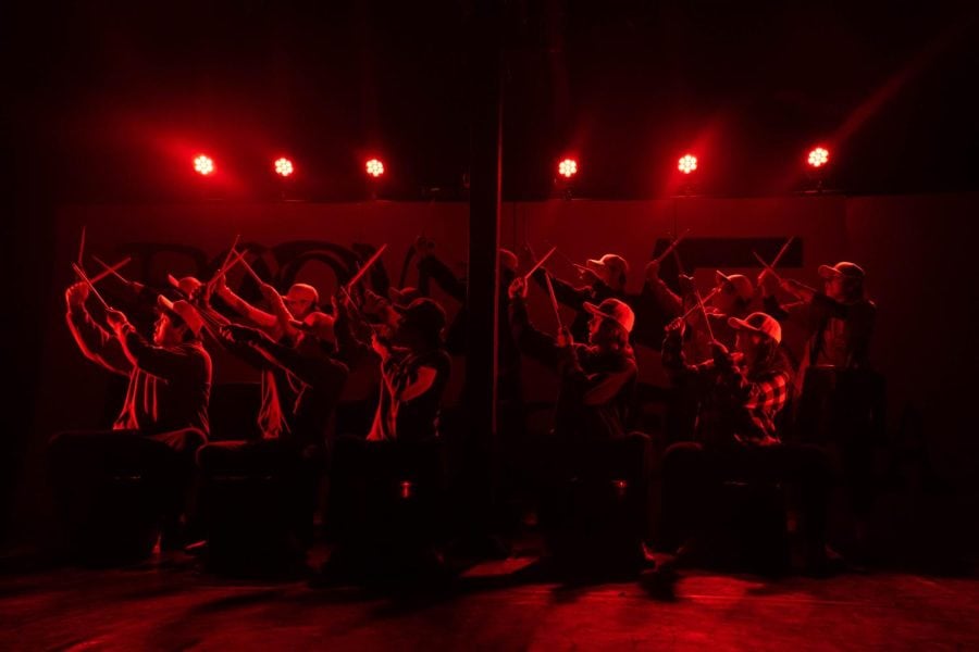 Several drummers tap their sticks together in a dark room with red lights.