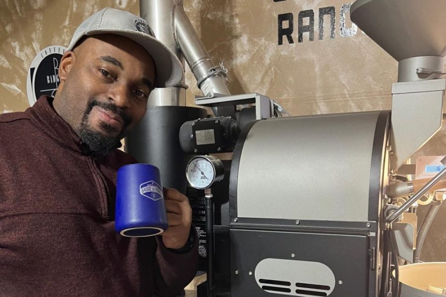 Mike Jones stands next to a coffee roaster in his garage with a cup labelled Badge Brew.