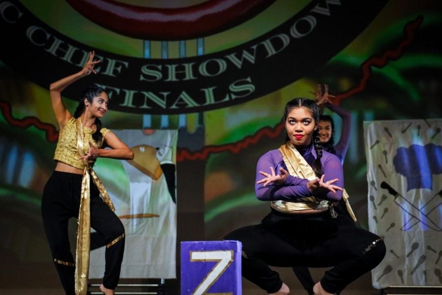 Two people dance on stage. One is in the foreground squatting and dressed in purple, while the other is dressed in gold and strikes a pose in the background.