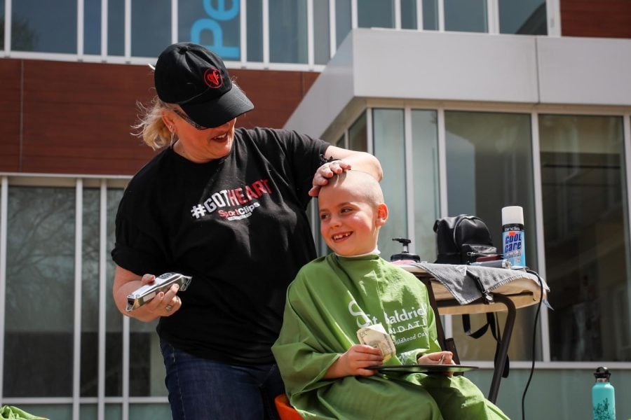 A little girl sits in a barber seat outside and gets her head shaved.