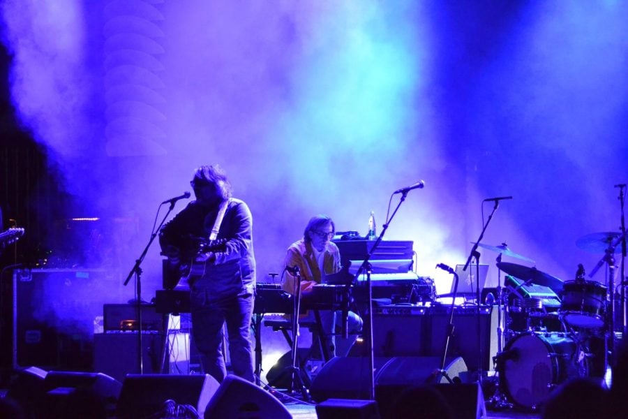Blue and purple lights and smoke fill the stage. Multiple microphones and drums are visible. A man with long hair, glasses, and a guitar is singing. There is a man with an orange jacket, seated playing keyboard.