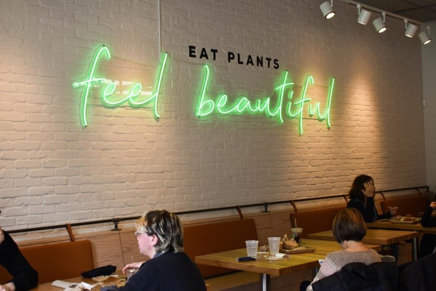 A white wall with black letters reading “EAT PLANTS” and a green neon sign reading “feel beautiful.” There are wooden booths and dining tables with patrons.