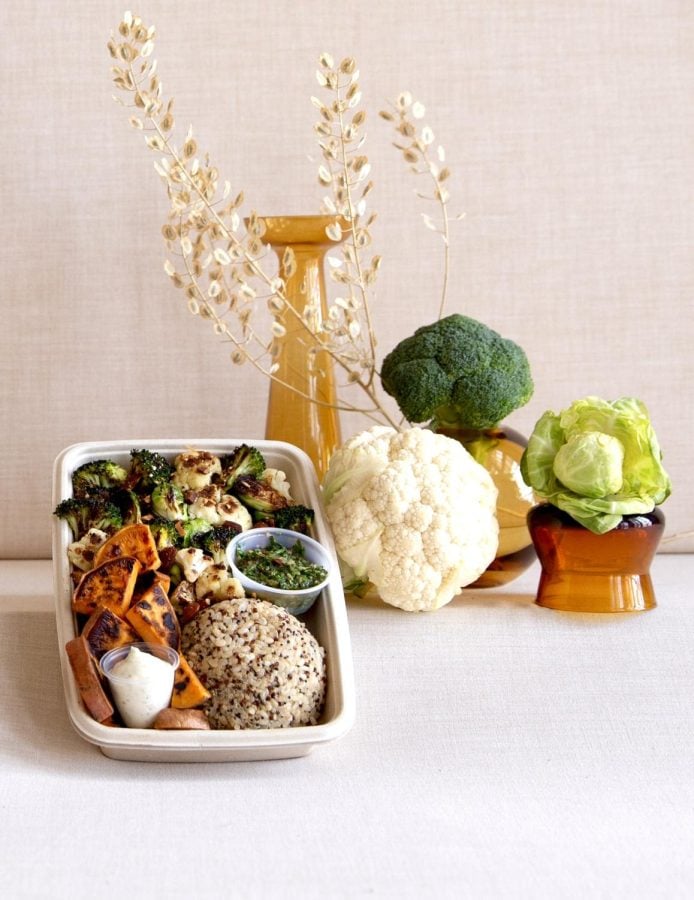 A container of food on the left with a pile of rice, vegetables and sauce containers. On the right is a pile of vegetables with a large piece of wispy dried grass.