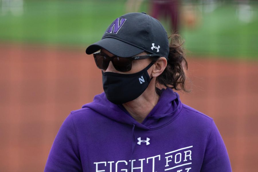 Softball coach in purple sweatshirt, hat, mask and sunglasses stands on field.