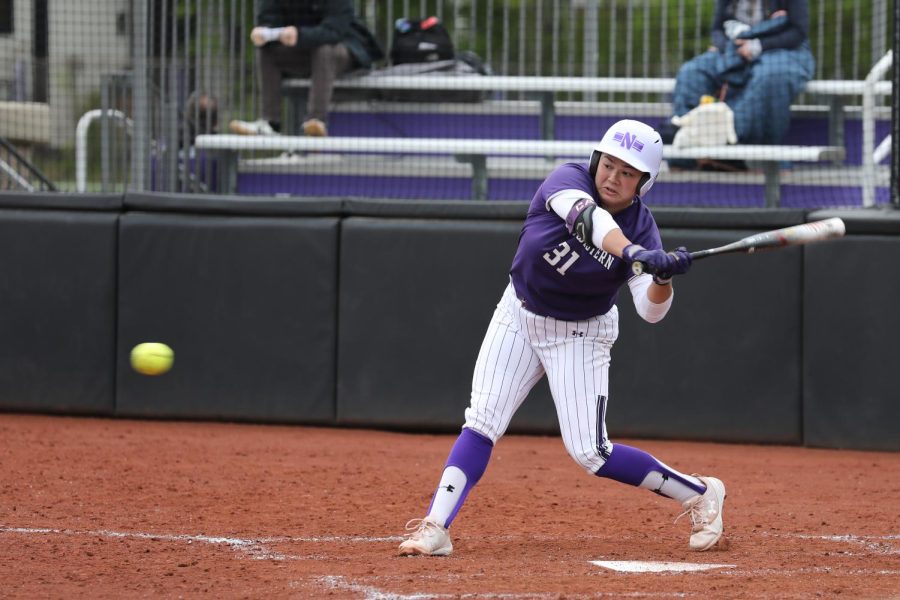 Softball player in purple-and-white uniform runs swings bat at incoming pitch.
