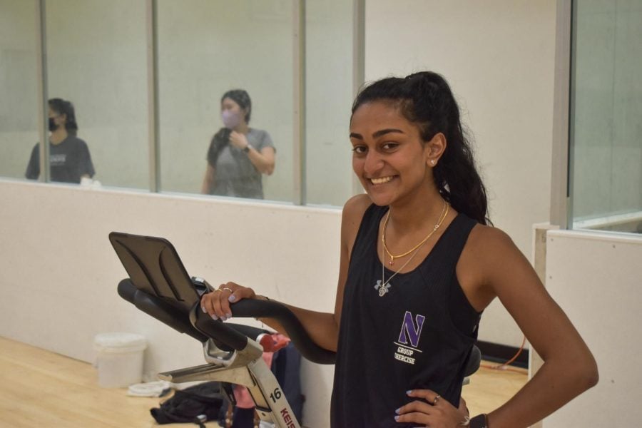 A student in a black tank top smiles at the camera with her hand resting on a stationary bike in the studio.
