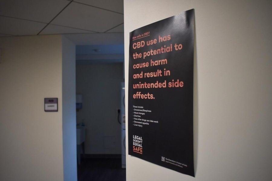 A black poster on a white wall: “How safe is CBD? CBD has the potential to cause harm and result in unintended side effects” in large orange letters. Below, it lists: “These include: Drowsiness/Sleepiness, Mood changes, Diarrhea, How other drugs you take work, Decreased appetite, Liver injury.” At the bottom, “LEGAL DOESN’T EQUAL SAFE” is written in capital letters.