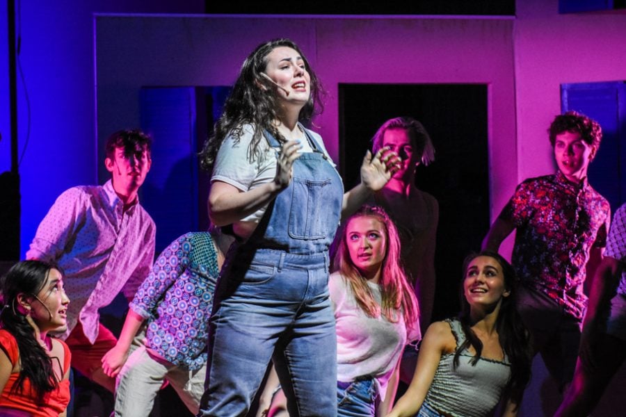 Actors are singing on a stage. At the center is an actor standing in blue overalls, and the rest of the actors look at her while she sings.