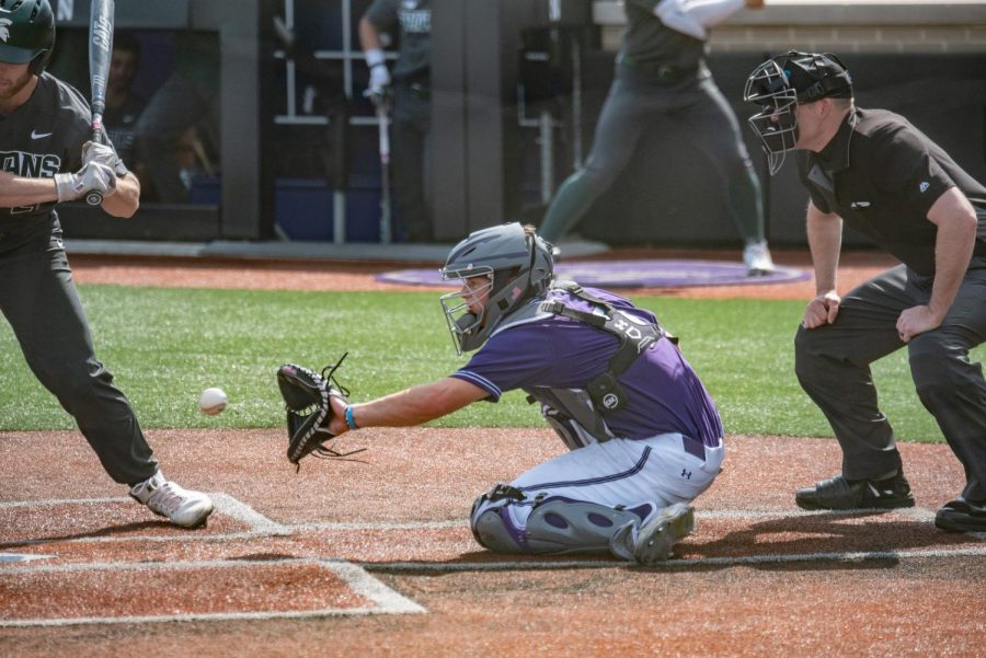 A catcher wearing a purple jersey, a helmet, and a chest protector catches a baseball.