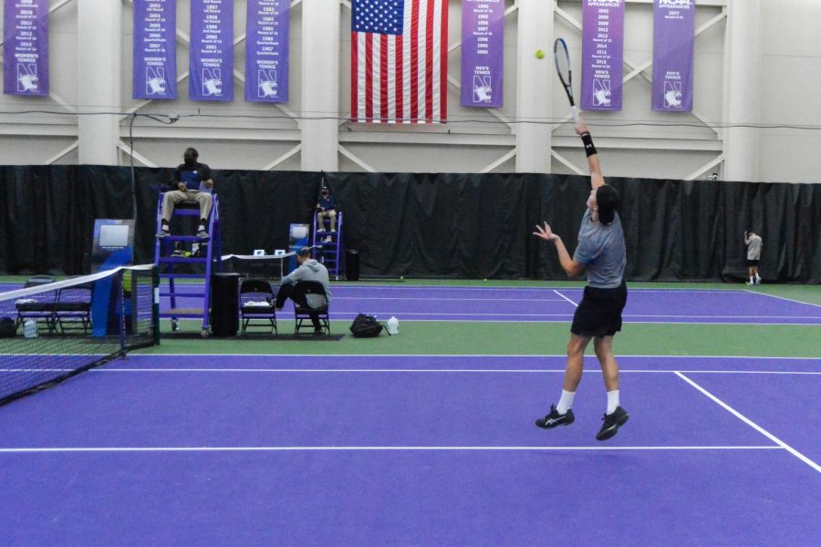 Male tennis player wearing gray shirt and black shorts holds racquet over his head.