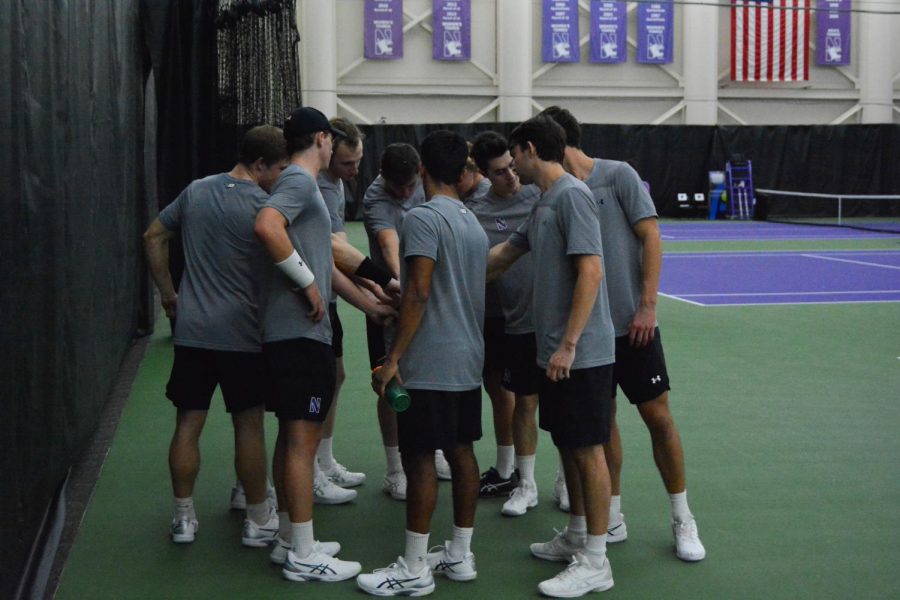 Tennis players in gray shirts and black shorts huddle on green and purple court.