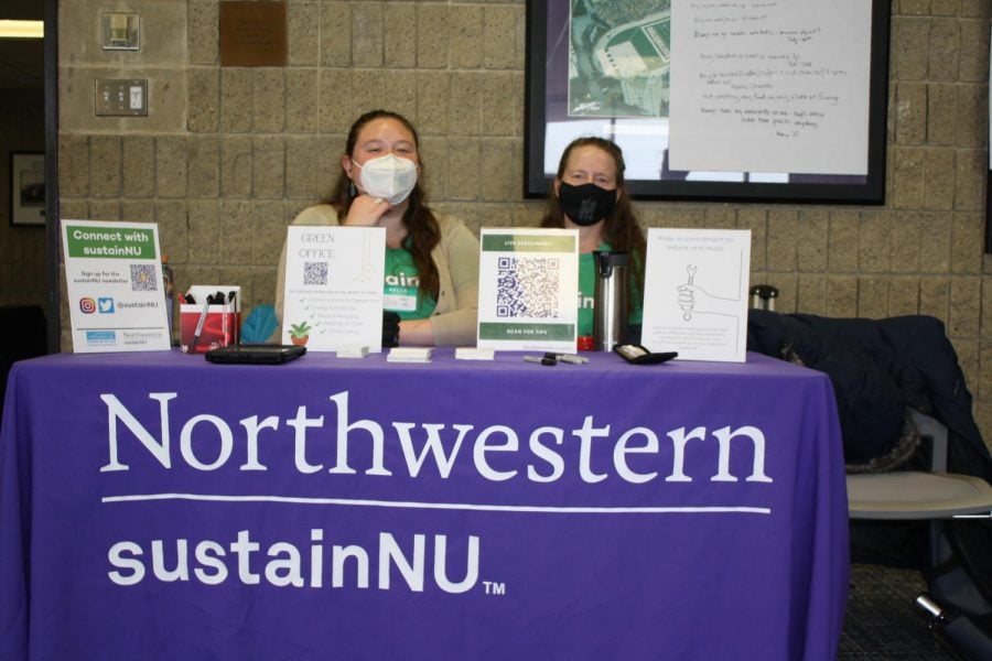 Two women sit at a purple table, in front of a gray brick wall that advertises sustainNU.